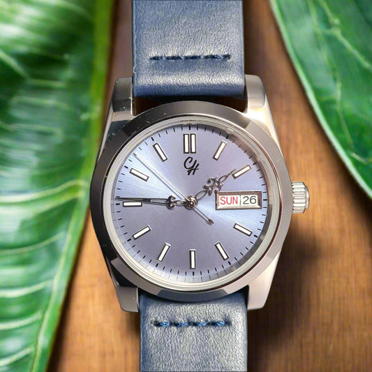 Wristwatch with blue leather strap on a wooden surface with green leaves