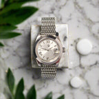 Silver watch with a mesh band on a marble surface with green leaves in the background